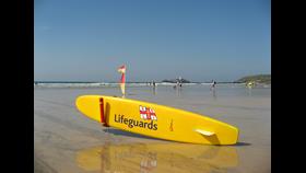 A yellow surf board is placed on the beach, it has the word lifeguards on it as well as the RNLI symbol.