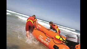 Two people guide a small orange boat into the water. The boat says 'Beach lifeguards' and has the RNLI symbol next to it.