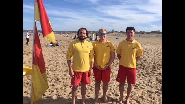Three RNLI lifeguards stand on a sandy beach. All are wearing yellow tops with RNLI on them and red shorts. Next to them are red and yellow flags.