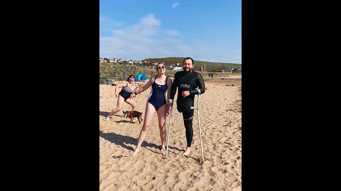 A man with an amputated leg stands on the beach with crutches next to a woman in a bathing suit.