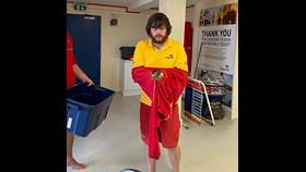 A RNLI lifeguard in a yellow top and red shorts is holding a small puffin in a red jumper. Besides him, a man is holding a container.