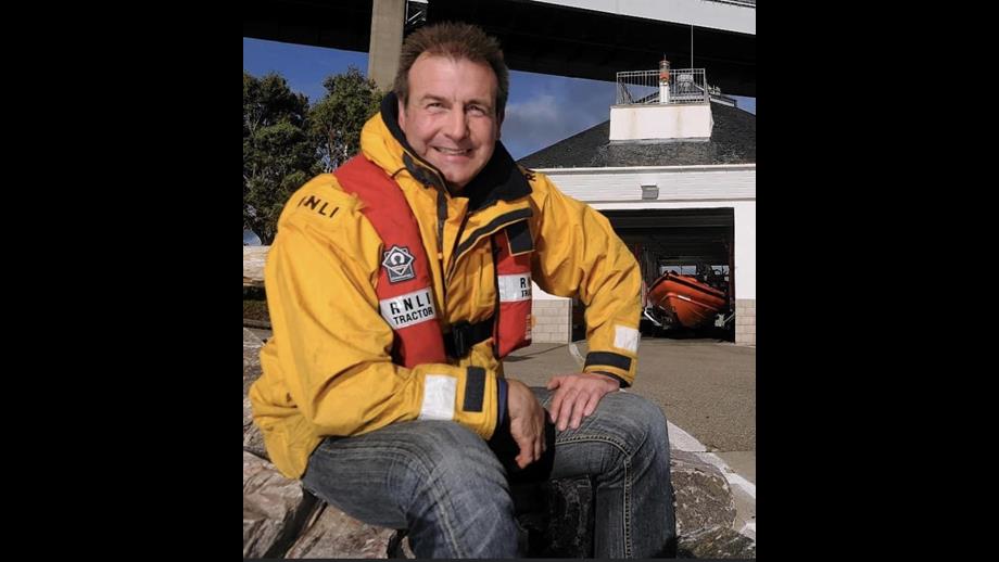A younger Alan sat outside the lifeboat station with the boat in the background inside he boat shed
