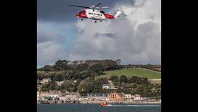 With Padstow the backdrop a volunteer crew member is extracted from Rocks D Class inshore lifeboat on a winch. The are member is photographed hanging mid air between the boat and the helicopter above.