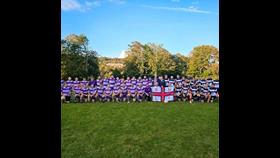 A photo of the Home Office Corsairs team members to the left, and RNLI rugby team members to the right, on a field in front of a rugby goal post. The people in the centre are holding an RNLI flag.
