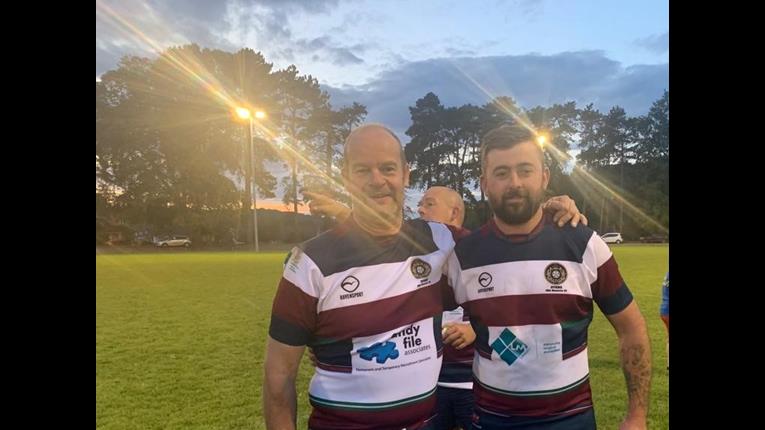 Two RNLI rugby team members are stood on a rugby pitch with their arm around eachother. They are wearing striped tops, and the floodlights are behind them.