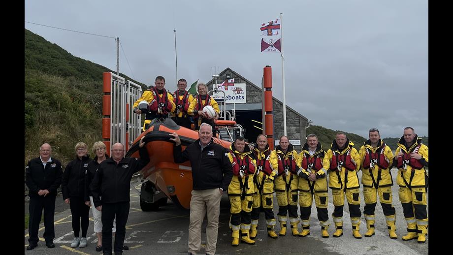 Line up of crew in front of Clifden lifeboat station. The inshore lifeboat is on a cradle in the background with three crew in drysuits standing. To the left of the photo are four volunteers lined up with another eight crew lined up on the right hand side of the lifeboat in full kit.