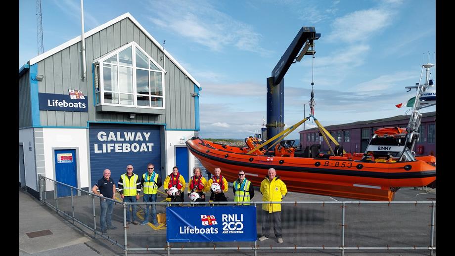 Eight crew lined up outside the Galway lifeboat station with the inshore lifeboat in the background to the right, held in position by the davit crane