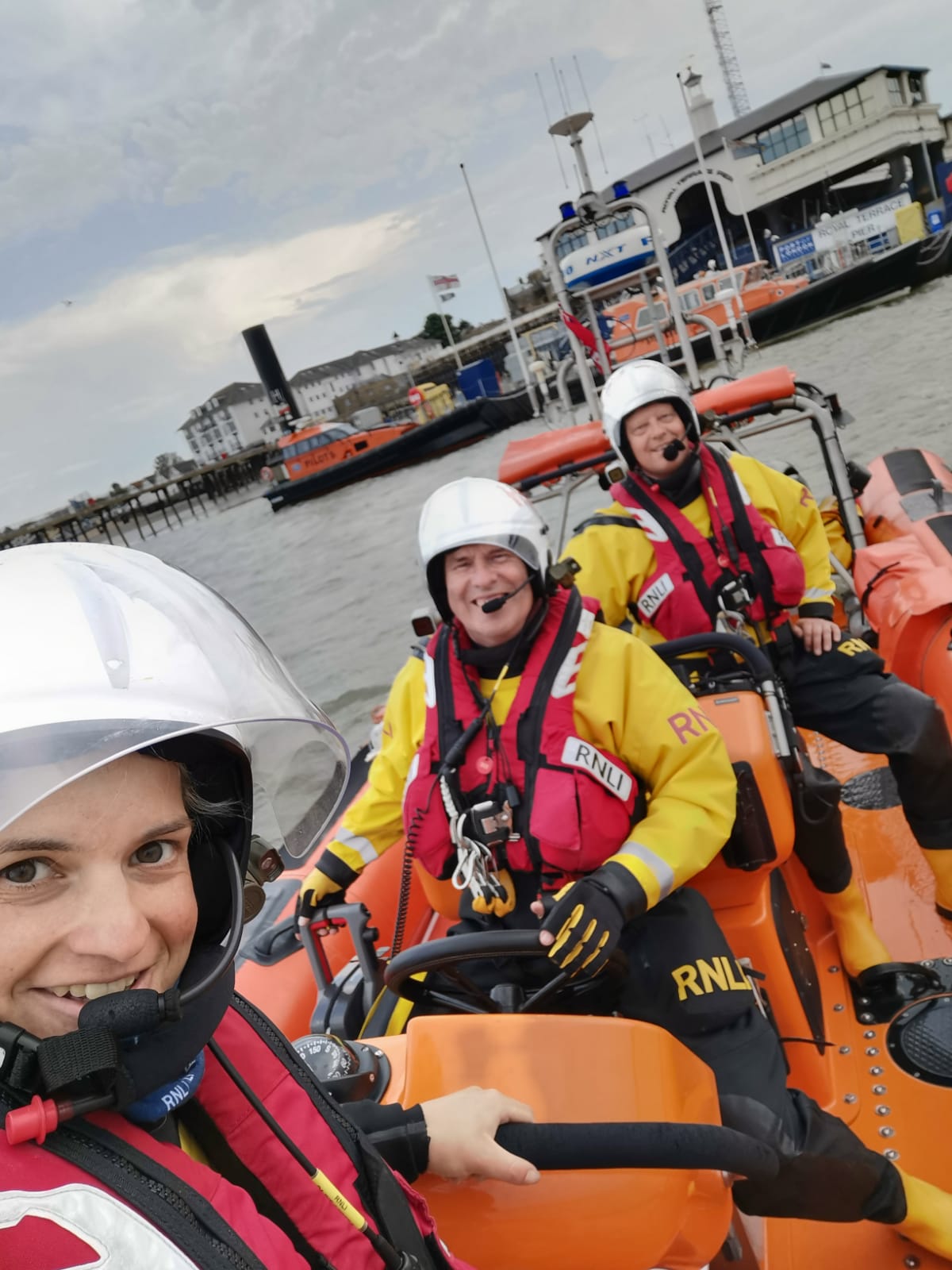 Three RNLI lifeboat crew members are pictured on a boat. A female stands at the helm, and two males sat further back. The crew is wearing an inshore lifeboat crew kit: a yellow drysuit, and red lifejacket. They are on an orange B class RNLI lifeboat, and smile at the camera. 