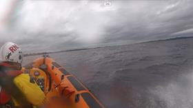 Looking over the right shoulder of the helm and starboard side of the lifeboat, is the rough gray lake, shoreline and the casualty vessel in the distance