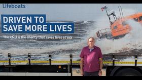 Charlie Davies, the recipient of a 50 year long service medal, stands beside an RNLI delivery lorry with the words driven to Save More Lives