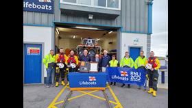 Ten crew standing outside the lifeboat station in Galway with the shutter door to the boat hall open and the inshore lifeboat just visible. In front there is a table with a lifeboats-branded tablecloth and on the table the case holding the scroll which is being held towards the camera by two crew in the centre. Three crew to the right are holding an RNLI200 banner.