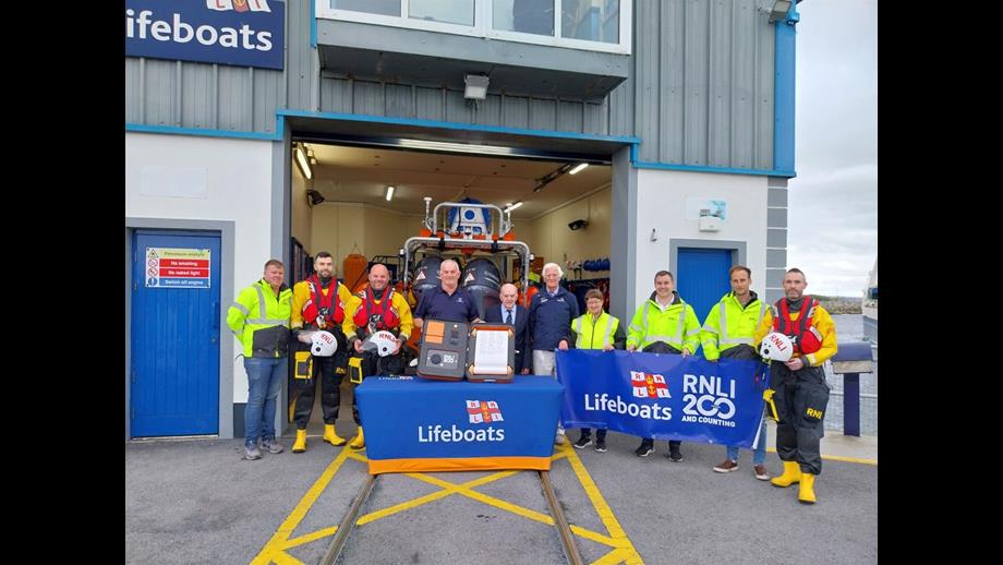 Ten crew standing outside the lifeboat station in Galway with the shutter door to the boat hall open and the inshore lifeboat just visible. In front there is a table with a lifeboats-branded tablecloth and on the table the case holding the scroll which is being held towards the camera by two crew in the centre. Three crew to the right are holding an RNLI200 banner.