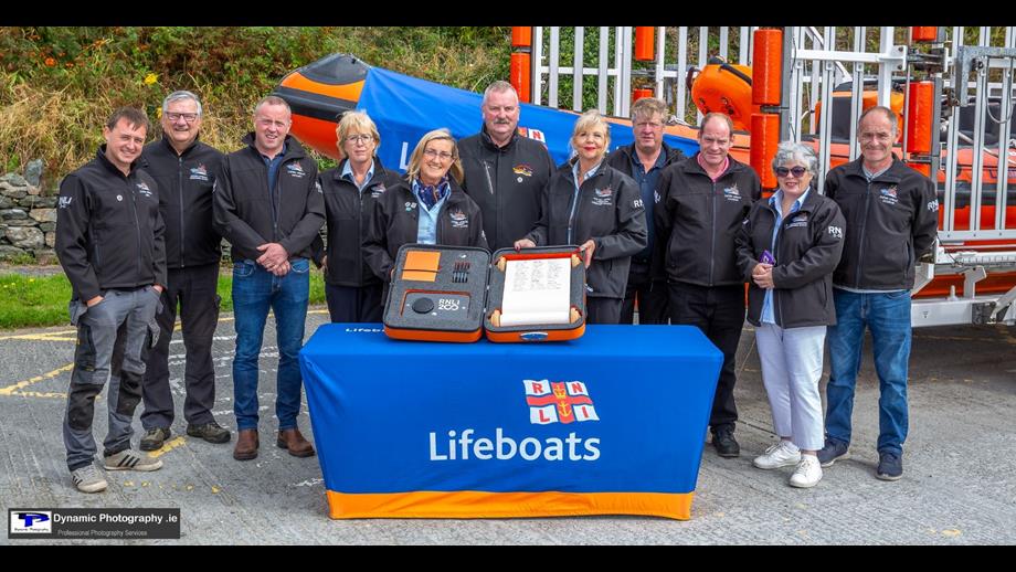 Eleven crew standing in a line outside with an inshore lifeboat on a trailer in the background. In front there is a table with a lifeboats-branded tablecloth and on the table the case holding the scroll which is being held towards the camera by three crew in the centre.