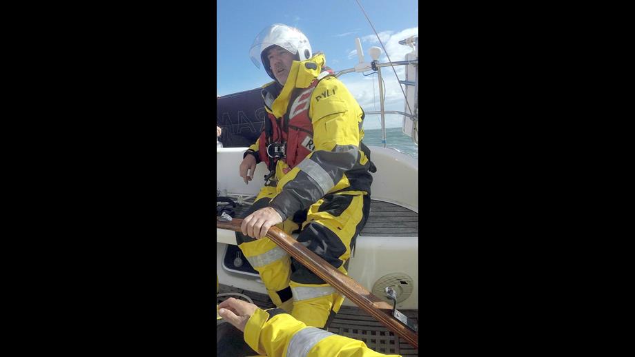 A Lifeboat crew member in full wet weather gear and lifejacket is at the tiller of a yacht