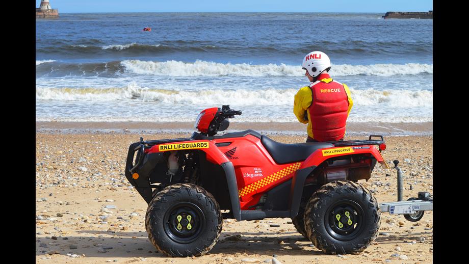Lifeguard leaning on the back of RNLI quadbike, with RNLI helmet on.
