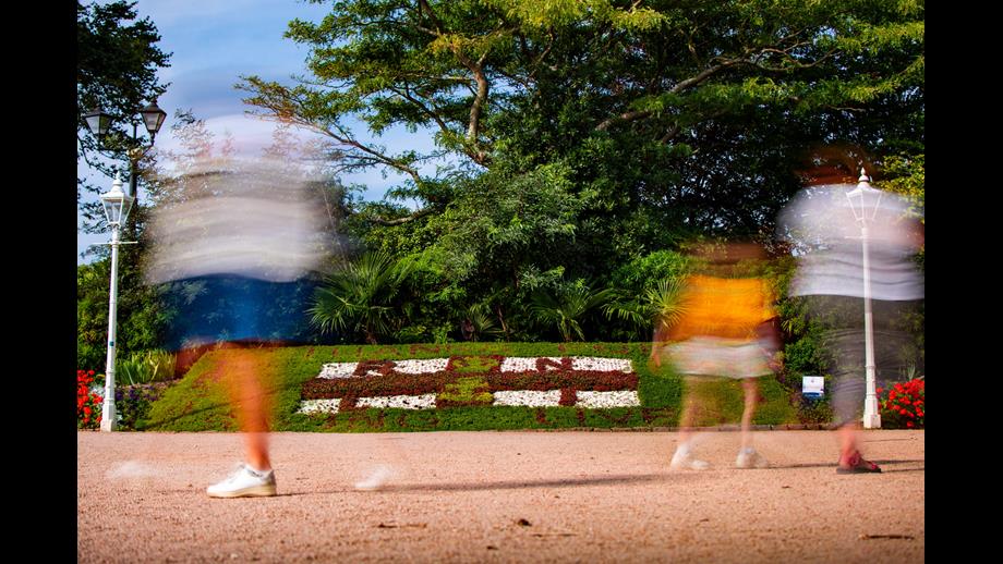A low level shot of the decorative flowerbed at Howard Davis Park, showing passers by in a blurred action shot in the foreground and the flowerbed in the background. The flowerbed is the RNLI flag with the words 'Thanks for' and 'Saving lives' at the top and bottom of the flag and '1824' to the left and '2024' to the right. 