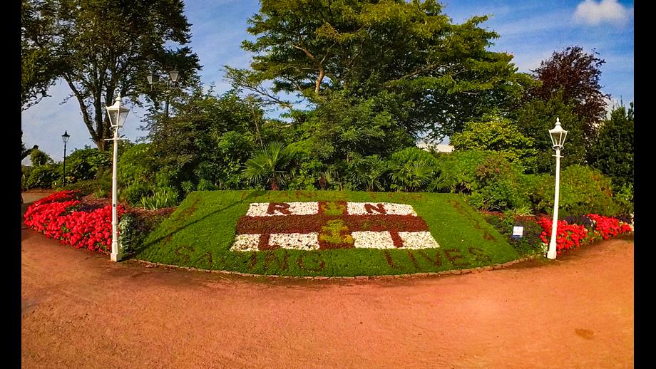 A wide angle shot of the decorative flowerbed at Howard Davis Park with the gravel path in the foreground and the flowerbed in the background under a blue sky and mature trees. The flowerbed is the RNLI flag with the words 'Thanks for' and 'Saving lives' at the top and bottom of the flag and '1824' to the left and '2024' to the right.