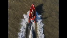 An orange lifeboat is central, viewed from above, in dark waters. The lifeboat has four crew in white helmets, yellow drysuits, and red lifejackets. There is wash coming from the back of the boat.