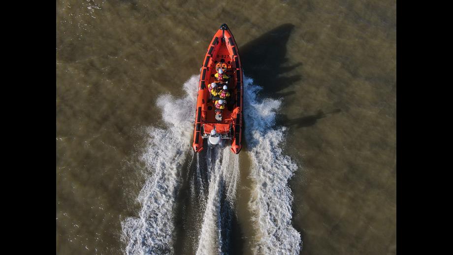 An orange lifeboat is central, viewed from above, in dark waters. The lifeboat has four crew in white helmets, yellow drysuits, and red lifejackets. There is wash coming from the back of the boat.