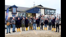 Press Gangers members stood in front of Aberdyfi Lifeboat Station with smiles and their fists in the air, alongside RNLI volunteer crew.