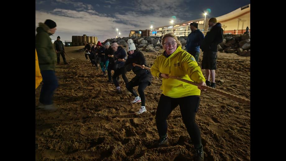 A night time shot, on New Brighton beach. On the beach in the foreground, a team of volunteers from New Brighton RNLI hold and pull hard on a long rope. Others stand nearby watching.