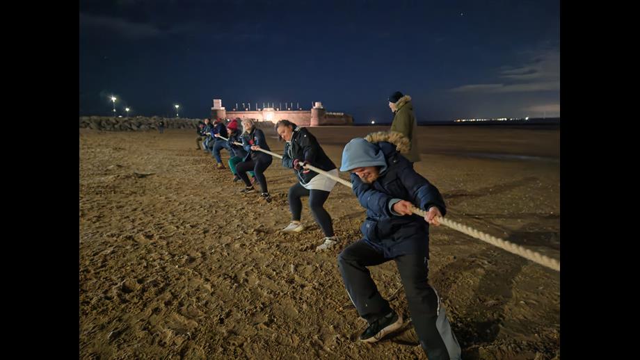 A night time shot, on New Brighton beach. On the beach in the foreground, a team of volunteers from New Brighton RNLI hold and pull hard on a long rope. Others stand nearby watching.