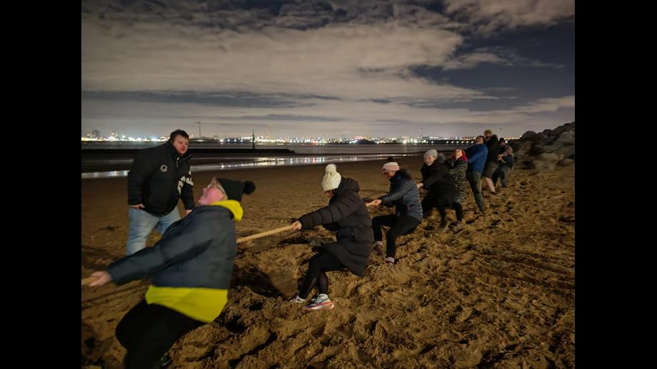 A night time shot, on New Brighton beach. On the beach in the foreground, a team of volunteers from New Brighton RNLI hold and pull hard on a long rope. Others stand nearby watching.