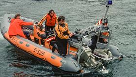 An orange and grey inflatable RNLI lifeboat, 'Lord Brotherton' (B-538), returns to its station on calm water. Four volunteer crew members in waterproof gear manage the boat, one operating controls, and others securing the vessel.