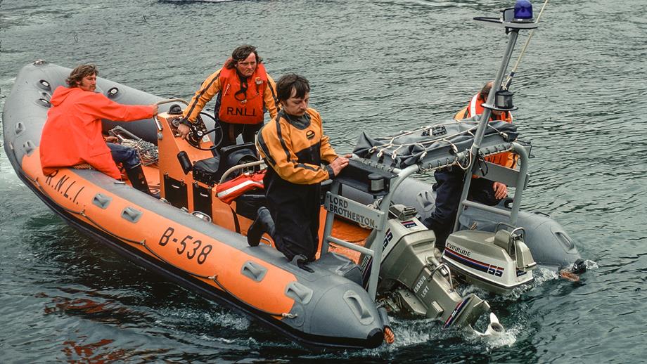 An orange and grey inflatable RNLI lifeboat, 'Lord Brotherton' (B-538), returns to its station on calm water. Four volunteer crew members in waterproof gear manage the boat, one operating controls, and others securing the vessel.