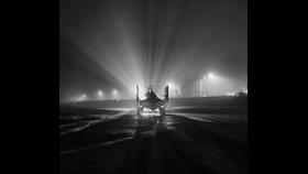 New Brighton RNLI's tractor, carrying the lifeboat, is heading toward the photographer, launching on service. In the fog, beams of light can be seen shining out from the lights behind into the sky. Black and white.
