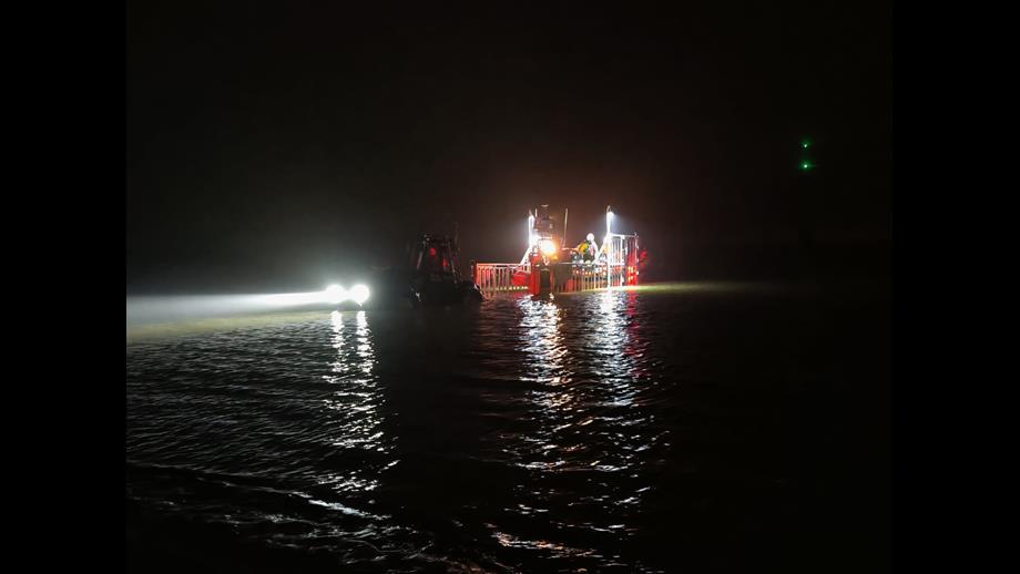 The lifeboat has recovered into the carriage in the dark. The river, sky and tractor can barely be seen, with light only coming from the tractor headlights and the carriage mast lights, illuminating the deck of the lifeboat.
