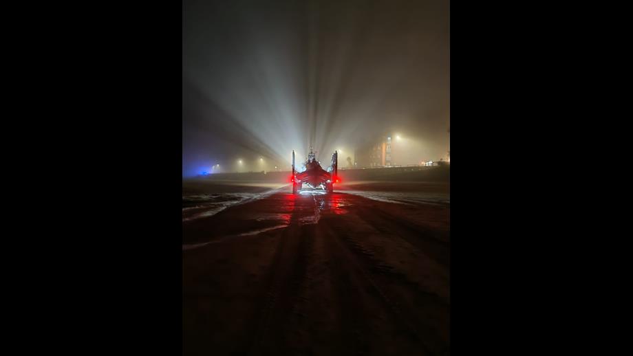 New Brighton RNLI's tractor, carrying the lifeboat, is heading toward the photographer, launching on service. In the fog, beams of light can be seen shining out from the lights behind into the sky. Colour image.