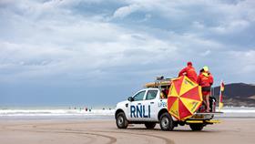 A white RNLI truck on a sandy beach carrying lifeguards wearing red and yellow and a sunshade