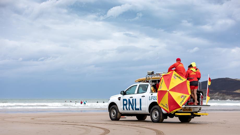 A white RNLI truck on a sandy beach carrying lifeguards wearing red and yellow and a sunshade