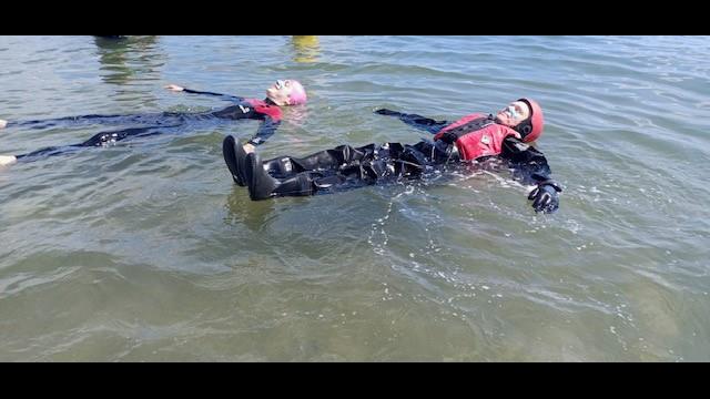 Two people, one in a drysuit and one in a wetsuit, float on their backs with their ears in the water and their arms outstretched