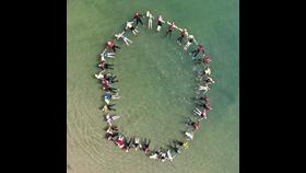 A birdseye view of 38 people connected in a circle floating on their back in the sea, limbs stretched out like a starfish.