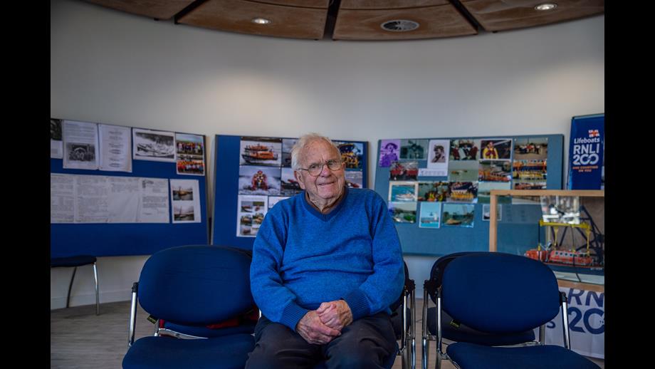 Man in blue jumper sitting on blue chair with photos on boards behind him 