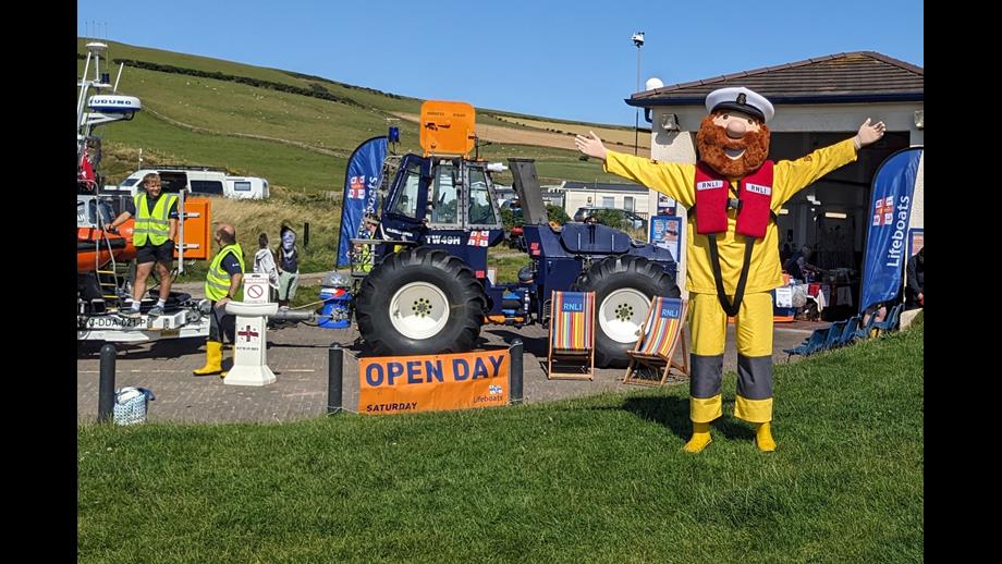 Stormy Stan the RNLI mascot has his arms open to welcome visitors standing in front of the tractor and lifeboat station