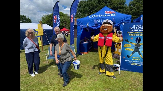 Fundraiser walking with collection bucket. RNLI Mascot Stormy Stand on the Right in front of blue gazebo