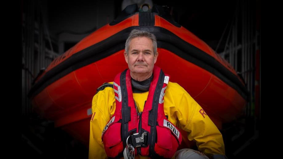 An RNLI crewmember in yellow drysuit and red lifejacket stands in front of an orange Atlantic 85 lifeboat