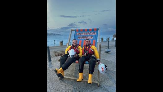 Two RNLI crew members in full kit sitting on an oversized deckchair on Folkestone Harbour Arm