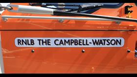 The name plate of Oban's new lifeboat with the words RNLB The Campbell-Watson in black on a white background on a panel against the orange upper structure of the Shannon class lifeboat.