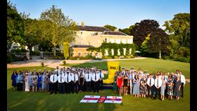 The RNLI Jersey crew, operations support, lifeguards and fund raisers gathered on the lawn of Government House in Jersey beside a giant yellow welly. Government House is in the background and an RNLI flag in the foreground. The sky is blue and everyone is dressed in their finery, with all the crew looking jolly smart in white shorts and RNLI ties.
