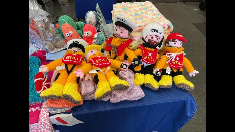 A craft table of knitted goods. Prominently featured are five knitted RNLI crew members in full PPE.
