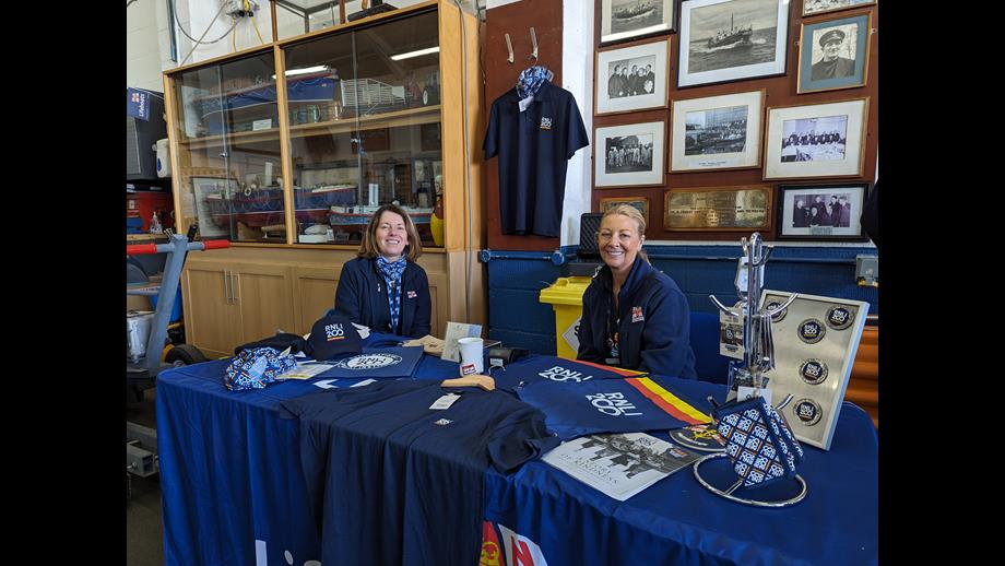 A stall inside New Brighton RNLI's boathouse features an array of 200th anniversary souvenirs. Two volunteers sit behind the table smiling at the camera.
