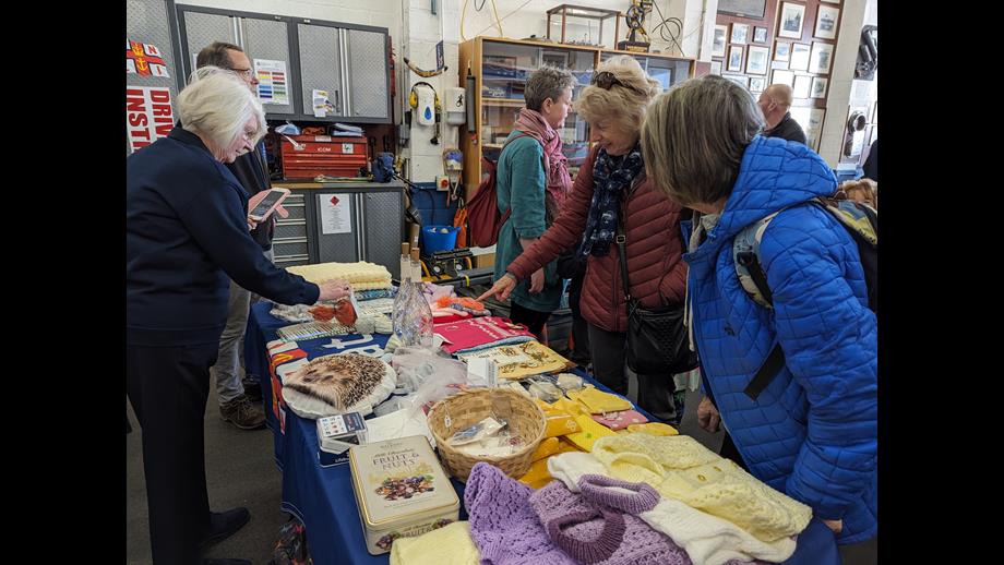 A stall inside New Brighton RNLI's boathouse displays an assortment of knitted goods. Two people are pointed excitedly toward items on the table.
