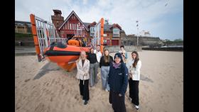 6 students and RNLI woman, infront of B-Class Lifeboat on sand outside of Cullercoats Lifeboat Station