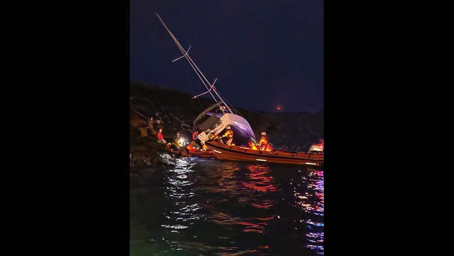 The stricken yacht lies across rocks in the background and the RNLI inshore lifeboats are in the foreground. It is dark, but the sea is calm.
