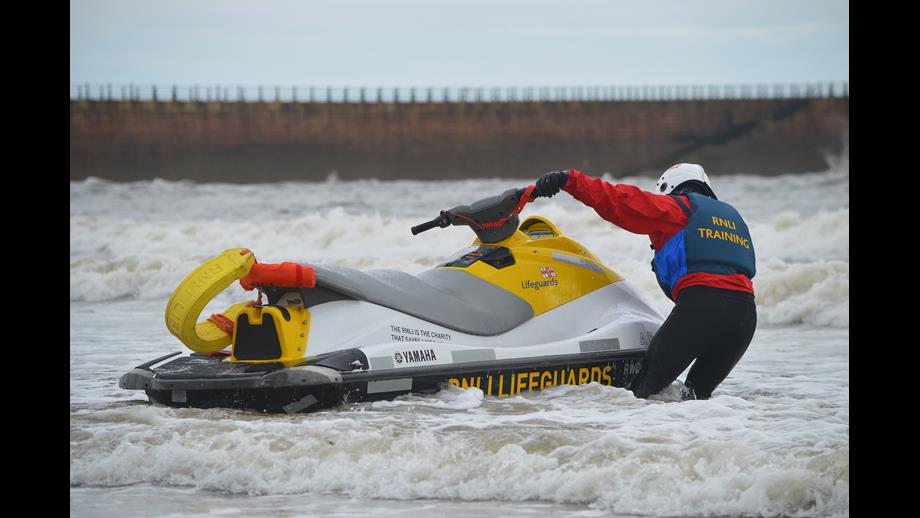 Man in wetsuit, lifejacket and helmet pulling a yellow jet ski through white water waves