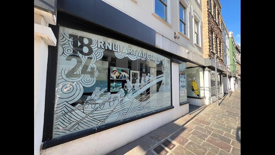 The decorated window display showing an etched outline of an Atlantic 85 perging into an old rowing lifeboat with the 1824 date and the words &quot;RNLI Art Auction&quot;. The paintings for auction are behind the etched window.